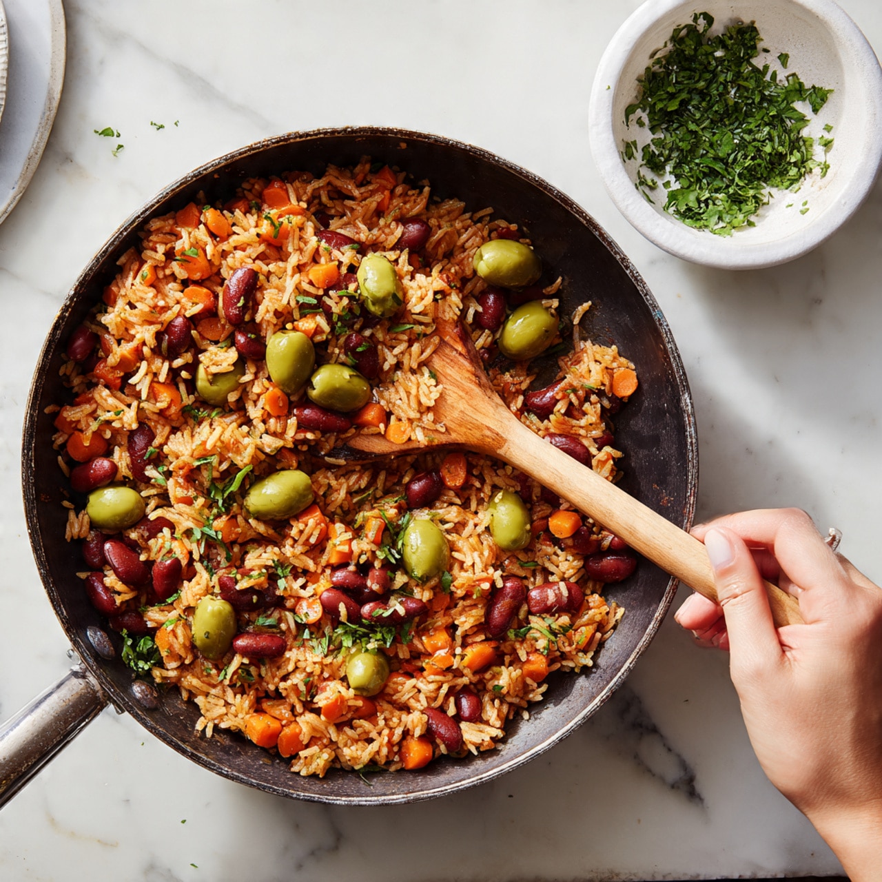A dark skillet filled with a cooked rice dish mixed with reddish-brown beans, sliced green olives, and small diced orange carrots, all evenly distributed in the dish. The rice looks moist and slightly glossy, with tiny green herb leaves sprinkled throughout. A woman's hand holding a wooden spatula is stirring some of the food from the bottom left corner of the skillet. The skillet is placed on a white marbled surface with a small round white bowl with herbs partially visible at the top right corner. Photo taken with an iphone --ar 4:5 --v 7