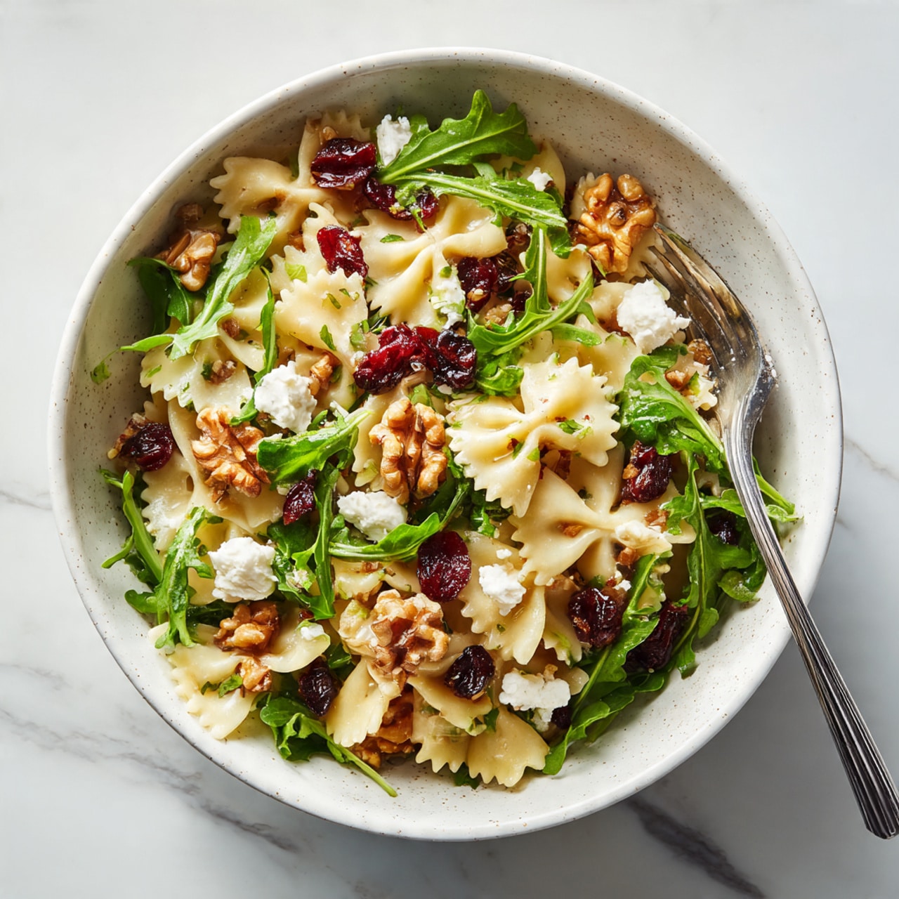 A white speckled bowl holds a salad with three main layers. The bottom layer is fresh, bright green leafy arugula, providing a leafy texture throughout. Over this are light brown farfalle pasta pieces, soft and slightly shiny from dressing, spread evenly across the greens. Scattered on top are medium brown walnut pieces with rough textures and some small creamy white cheese chunks. Dark red dried cranberries are mixed throughout, adding a pop of color. A silver fork rests in the bowl on the right side. The bowl sits on a white marbled surface. Photo taken with an iphone --ar 4:5 --v 7