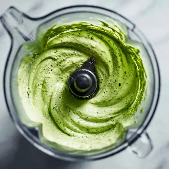 In the image, there is a clear blender jar filled with a thick, creamy green sauce that has a smooth texture with small bits of darker green herbs mixed in. The sauce creates soft waves and swirls around the central black blender blade, showing its thick consistency. The blender jar is resting on a white marbled surface. photo taken with an iphone --ar 4:5 --v 7