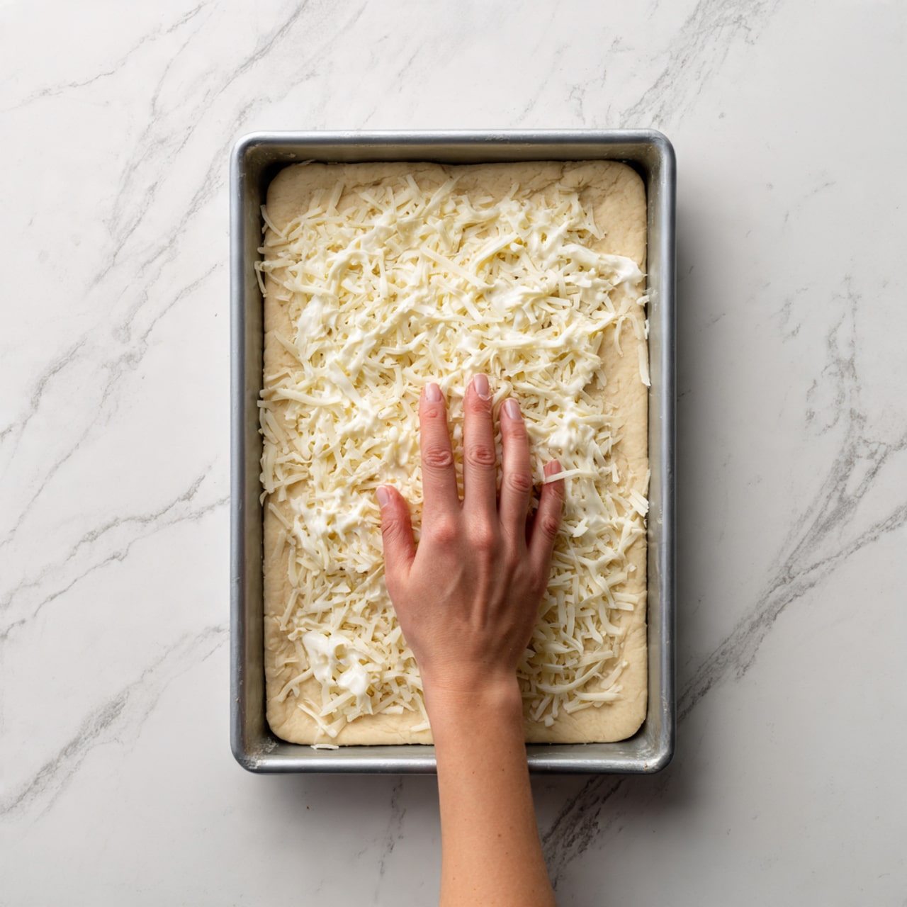 A close-up view of a rectangular metal baking tray filled with an uncooked pizza. The bottom layer is a smooth, pale dough evenly spread across the tray. On top of the dough is a light, even layer of white sauce that covers the entire surface. The topmost layer is scattered shredded white cheese spread unevenly over the sauce, showing patches of the sauce beneath. A woman's hand is visible on the right side, holding the edge of the tray. The tray rests on a white marbled surface. Photo taken with an iphone --ar 4:5 --v 7