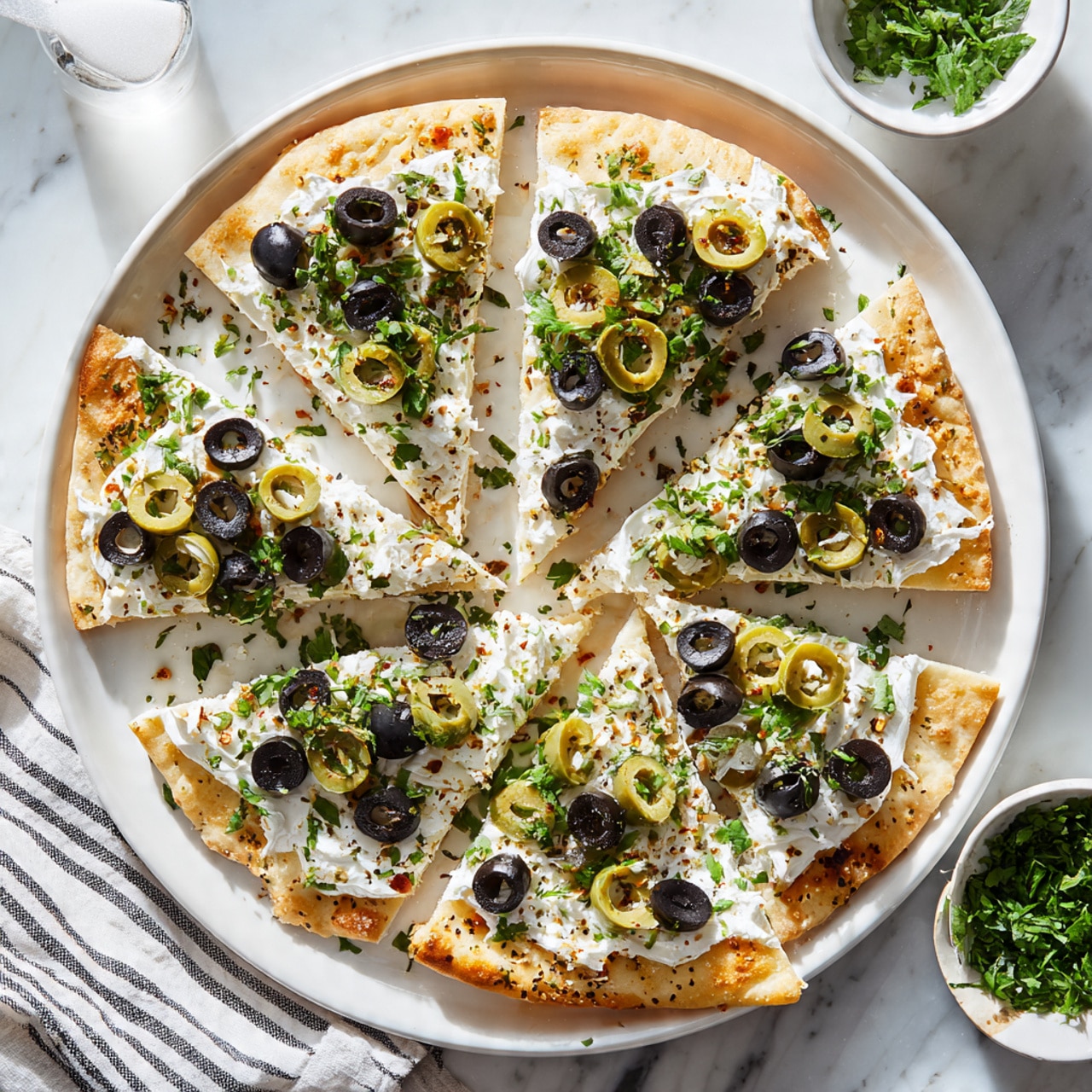 The image shows a white baking tray with a flatbread cut into several irregular triangles, each piece topped with a thick, creamy white cheese layer. The cheese is speckled with sliced black olives and green olives stuffed with small red pieces, evenly scattered on top. Bits of chopped fresh green herbs are sprinkled over the flatbread for a pop of color. On the upper right side of the tray, there's a small white bowl filled with finely chopped green herbs. The tray is placed on a white marbled surface next to a striped cloth. Photo taken with an iphone --ar 4:5 --v 7