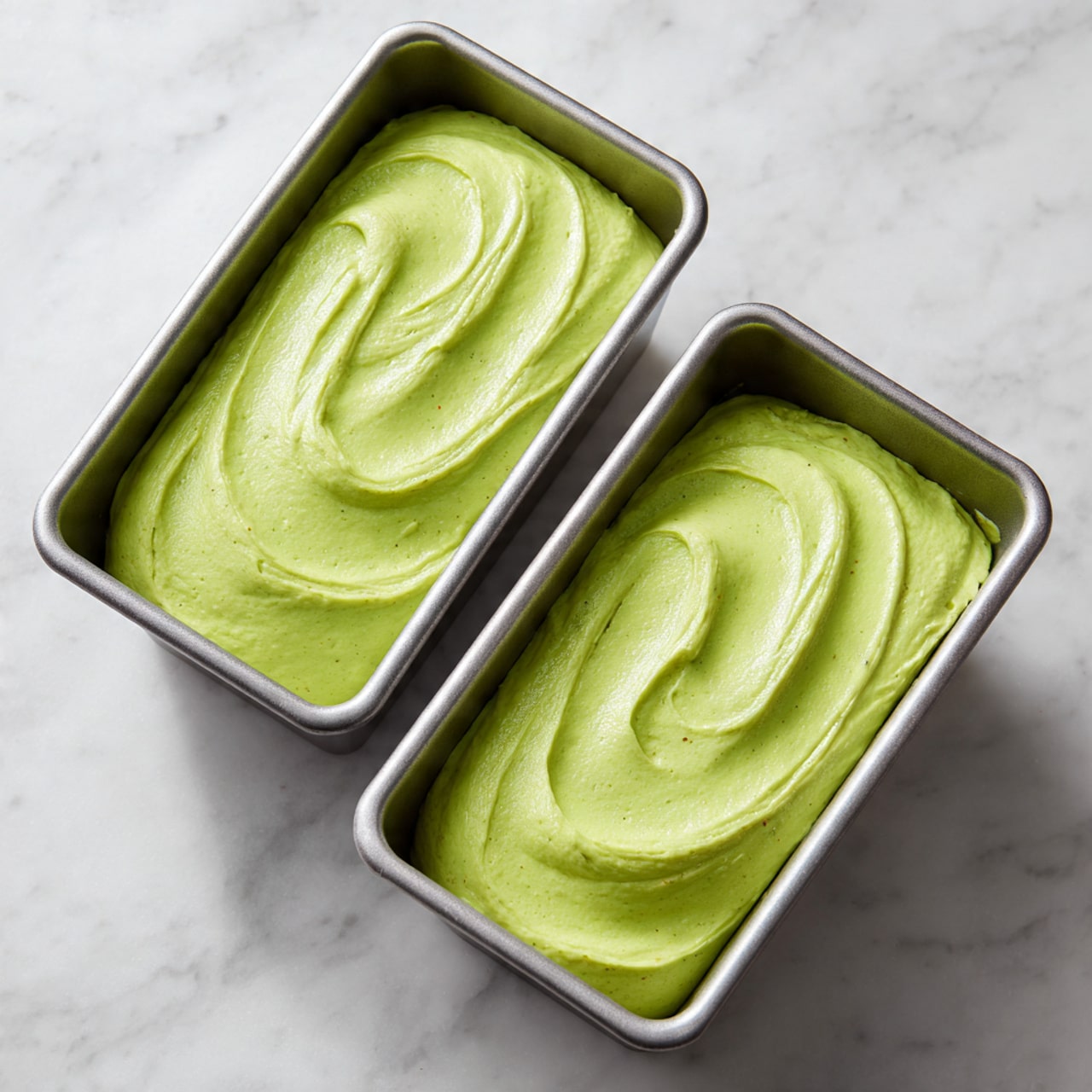 Two rectangular silver baking pans are filled with a smooth, light green batter, spread evenly across each pan. The batter has a soft texture with a few small brownish specks visible throughout. The pans sit on a white marbled surface, and the top of the batter has subtle swirls and lines from being spread. Photo taken with an iphone --ar 4:5 --v 7