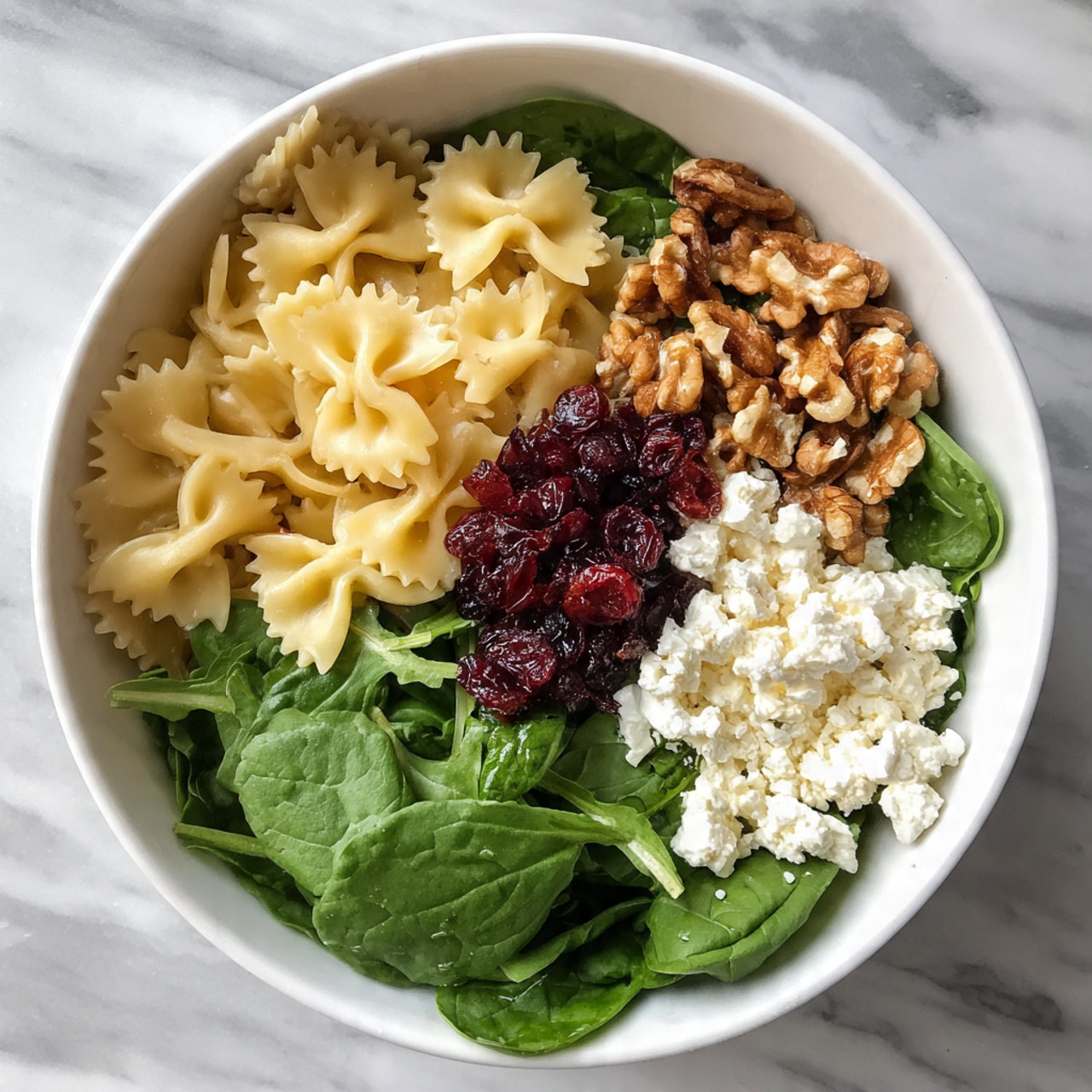 A white bowl filled with a colorful salad arranged in sections. One section has light yellow bowtie pasta with a smooth texture, next to it is a pile of small dark red dried cranberries with a slightly wrinkled look. Beside the cranberries, there is a crumbled white cheese with a soft and crumbly texture. In the middle, there is a bunch of brown walnuts with a rough and bumpy surface. On one side, bright green spinach leaves with visible veins and smooth surfaces, and on the other side, arugula leaves that are darker green with a more jagged shape. The bowl is placed on a white marbled surface. photo taken with an iphone --ar 4:5 --v 7