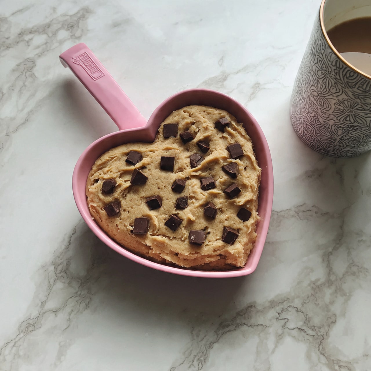 A heart-shaped pan with a pink handle is filled with thick, light yellow cookie dough, spread evenly to cover the pan's bottom and sides. Mixed throughout the dough are numerous deep brown chocolate chunks of various sizes, some square and some irregular, embedded in the dough and slightly melting at the edges. The pan sits on a white marbled surface, and there is a glimpse of a white cup with a black pattern at the top right corner. Photo taken with an iphone --ar 4:5 --v 7