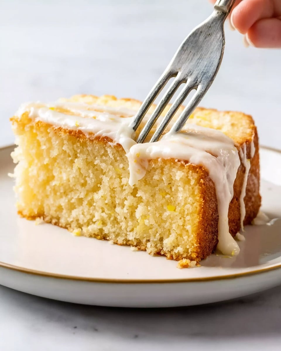 A close-up image of a single slice of yellow cake with a crumbly top layer and a smooth white icing dripping slightly down the sides. A silver fork is touching the cake from the top right corner, pressing gently into the cake, with a woman's hand holding the fork. The cake slice is on a simple white plate, placed on a white marbled surface. The textures show the softness of the cake and the creamy icing clearly. Photo taken with an iphone --ar 4:5 --v 7