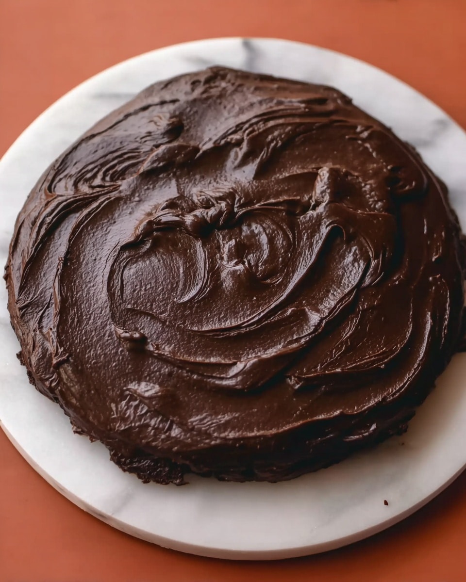 A round chocolate cake covered in thick, smooth, dark chocolate frosting with visible swirling patterns on the top. The cake surface has a glossy, creamy texture that suggests rich, dense chocolate. The cake is placed on a white plate sitting on a white marbled textured surface. Photo taken with an iphone --ar 4:5 --v 7