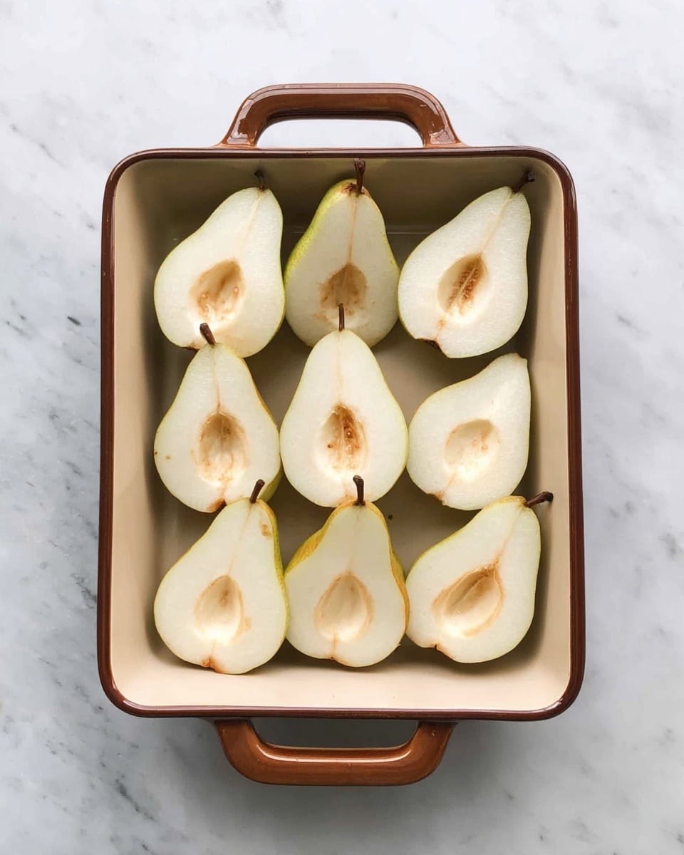 A rectangular baking dish with a brown rim and handles holds eight halved pears, arranged in two rows of four halves each, with the pear cores removed creating hollow centers; the pears show a smooth pale yellow color with slightly browned spots near the core area. The dish rests on a white marbled surface. photo taken with an iphone --ar 4:5 --v 7