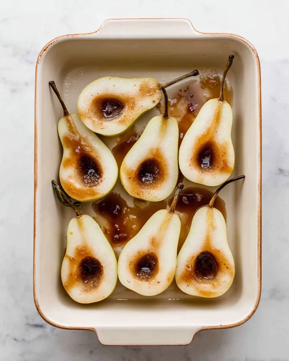 A white ceramic rectangular baking dish filled with eight halved pears, each pear half placed with the cut side up. The pears are pale yellow with a smooth texture, and each core area is hollowed out and filled with a dark brown syrup that has dripped slightly onto the baking dish surface. The stems remain on some pear halves, adding a natural touch. The baking dish sits on a white marbled surface. photo taken with an iphone --ar 4:5 --v 7