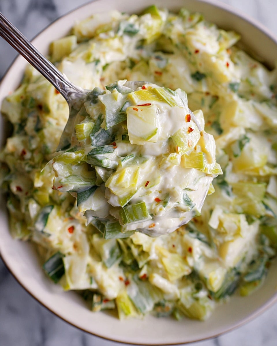 A close-up view of a creamy mixture with small diced pieces of light green and yellow vegetables in a white bowl, with the texture looking soft and slightly chunky; some white shreds are sprinkled on top, adding thin, fine texture. A silver spoon lifts a scoop of the creamy vegetable mix, showing layered bits of green, white, and yellow with a smooth white sauce holding them together, with tiny red flakes visible. The background is a white marbled surface. Photo taken with an iphone --ar 4:5 --v 7