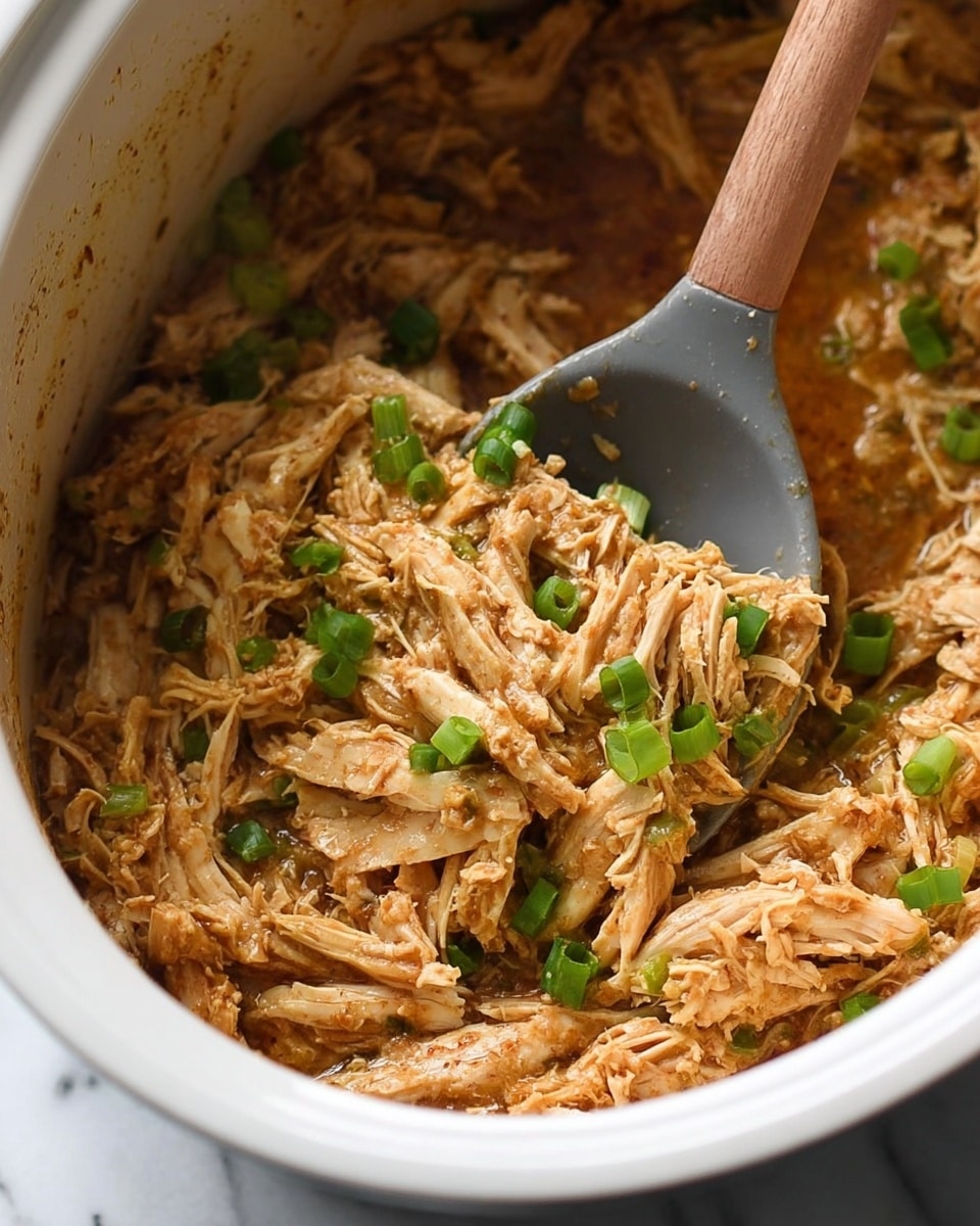 The image shows a close-up of shredded cooked chicken mixed with a light brown sauce and small green onion pieces scattered on top. The chicken is in a white pot, and a gray spoon with a wooden handle is scooping some of the chicken from the pot. The texture of the chicken looks moist and tender, layered densely with the sauce coating each shred evenly. The background is a white marbled surface. photo taken with an iphone --ar 4:5 --v 7
