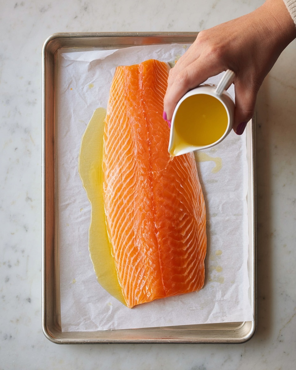 A large, raw salmon fillet lies flat on a sheet of white parchment paper atop a silver baking tray. The salmon is bright orange with fine white lines running evenly through it, showing its fresh texture. A woman's hand is pouring a small amount of golden oil from a white small pitcher onto the middle of the fish, creating a shiny wet trail on the surface. The baking tray sits on a white marbled surface, adding a clean and simple background to the scene. photo taken with an iphone --ar 4:5 --v 7