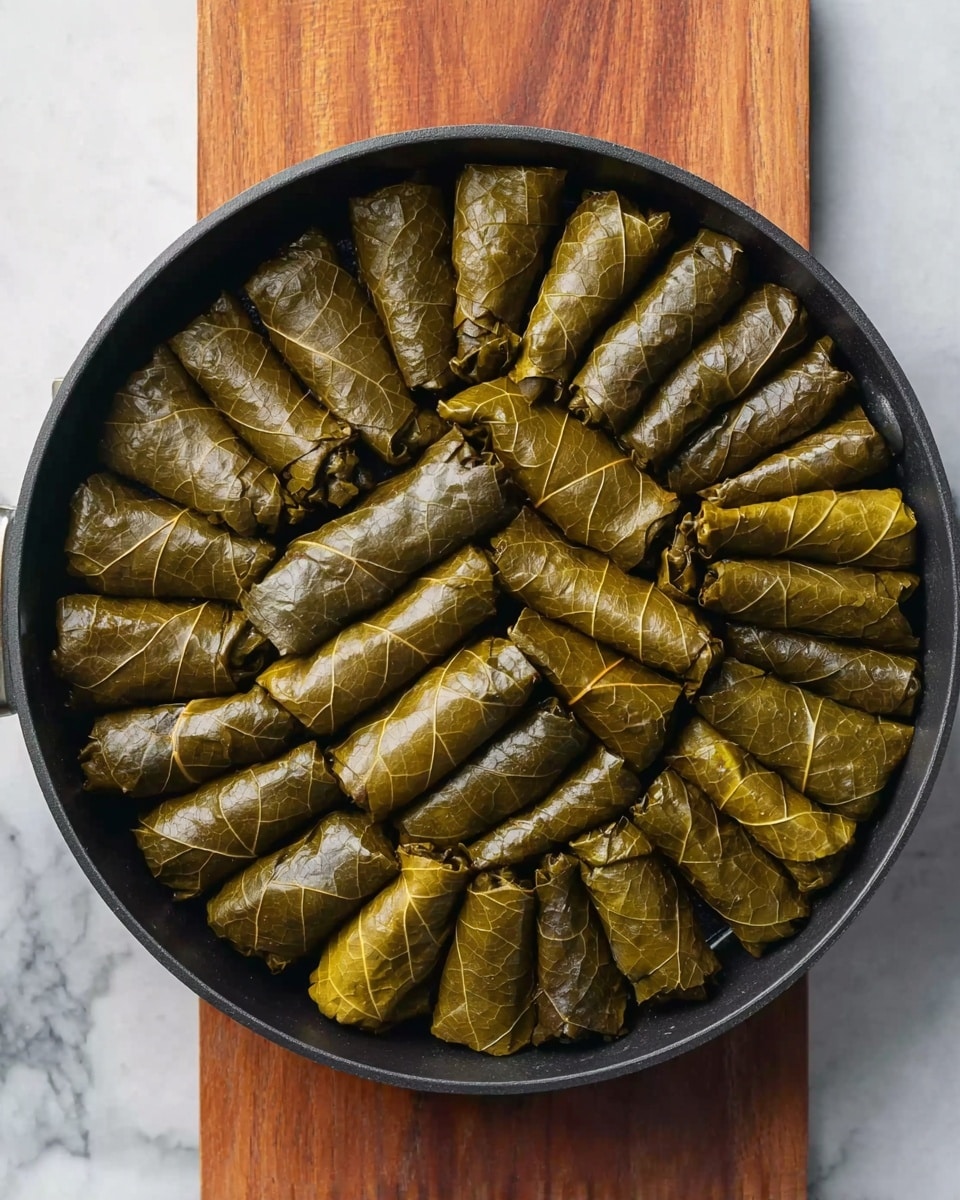 A round black pan filled with neatly arranged green grape leaves stuffed with a filling, each leaf rolled tightly into small, uniform cylindrical shapes. The grape leaves have a slightly shiny texture and vary in shades of green, from medium to dark. The pan is placed on a rectangular wooden board that rests on a white marbled surface. The grape leaves cover the entire pan, stacked closely together without visible layers underneath. Photo taken with an iphone --ar 4:5 --v 7