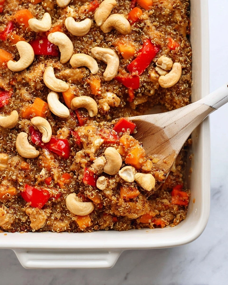 A white baking dish filled with a close-up view of a mixed quinoa casserole. The dish has three main layers: the base is a soft, brown mix of quinoa and sauce; scattered throughout are chunks of light brown cooked chicken and bright pieces of red and orange bell peppers; on top, there is a generous layer of whole light beige cashew nuts. A wooden spoon with a white handle is partly inside the dish, scooping some of the mixture from the bottom right corner. The background is a white marbled surface. photo taken with an iphone --ar 4:5 --v 7