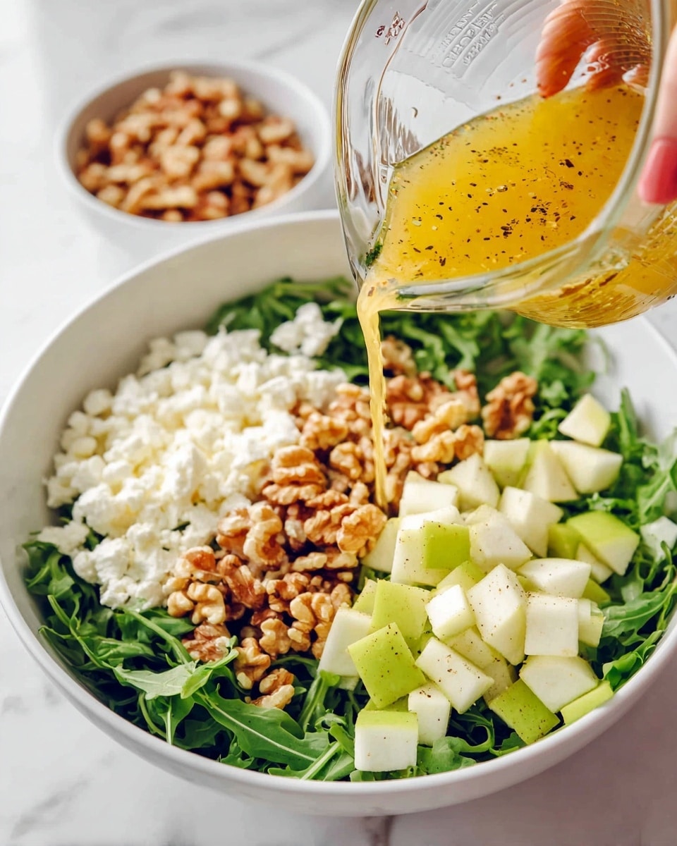 A white bowl filled with layers of fresh green arugula at the bottom, topped with three distinct sections: white crumbled cheese on the left, a pile of light brown walnut pieces in the center, and small cubes of green and white pear on the right. A clear glass measuring cup is held by a woman's hand above the bowl, pouring a golden-yellow dressing with visible black pepper flakes over the salad. In the background, a separate white bowl holds more walnut pieces. All this is placed on a white marbled surface. photo taken with an iphone --ar 4:5 --v 7