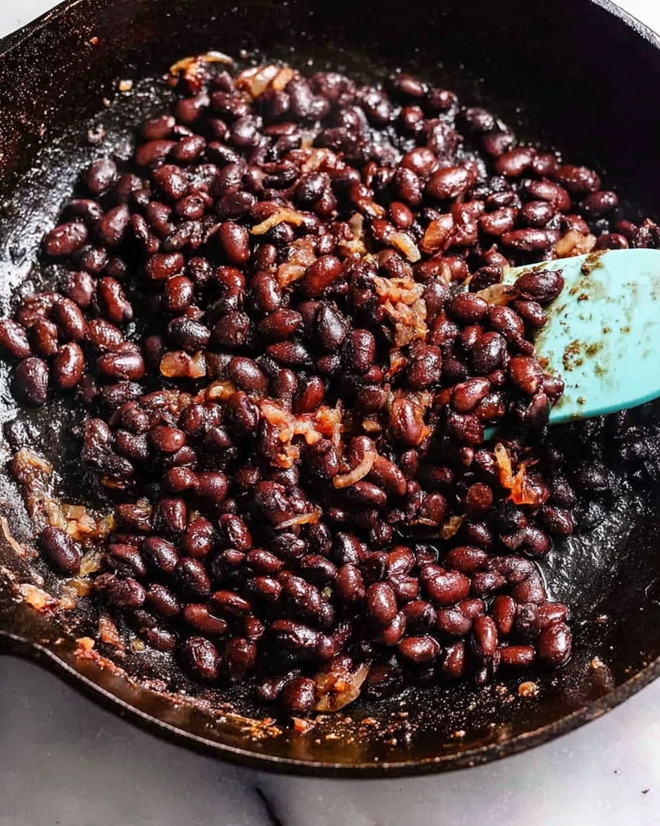 The image shows a close-up of black beans cooking in a black cast iron pan. The beans are dark brown to black in color with a shiny texture indicating they are soft and cooked. Some beans look slightly mashed and mixed with small bits of caramelized onions or spices. A light blue silicone spatula is partly visible on the right side of the pan, stirring the beans. The pan surface has a well-used, rustic look with some seasoning marks. The background is a white marbled texture. photo taken with an iphone --ar 4:5 --v 7