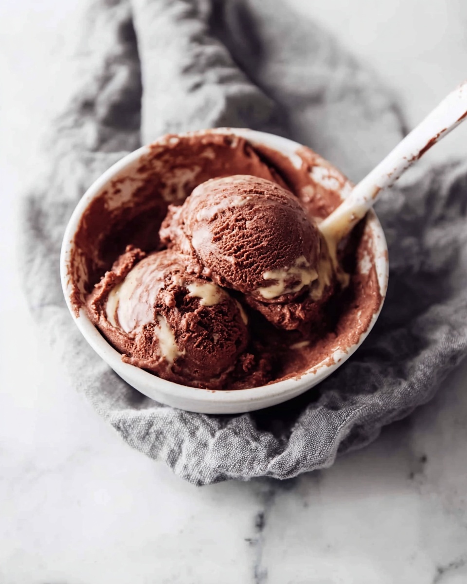 Two scoops of dark brown chocolate ice cream with lighter swirls of creamy beige sauce on top sit in a small white bowl. The texture looks soft and slightly rough. A white spoon with some chocolate smudges is placed inside the bowl on the right side. The bowl rests on a crumpled gray cloth over a white marbled surface. The photo taken with an iphone --ar 4:5 --v 7
