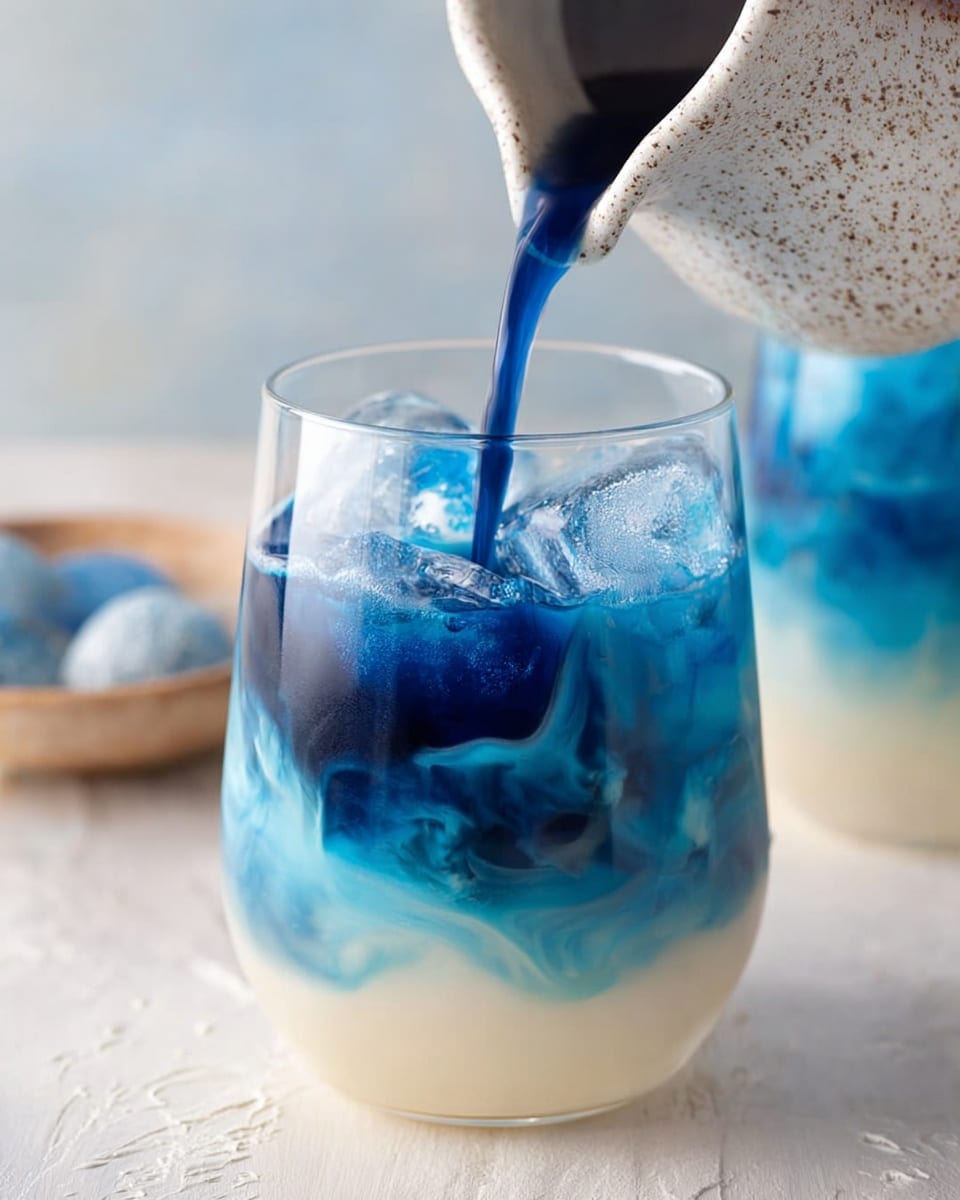 A clear glass holds a three-layer drink with a white creamy base filling about half the glass, a layer of swirling light blue above it blending into deeper blue tones with ice cubes visible in this middle section, and a dark blue liquid being poured from a speckled white ceramic pitcher into the glass from above, catching movement and mixing colors. The background shows another similar glass and objects on a white marbled textured surface. photo taken with an iphone --ar 4:5 --v 7