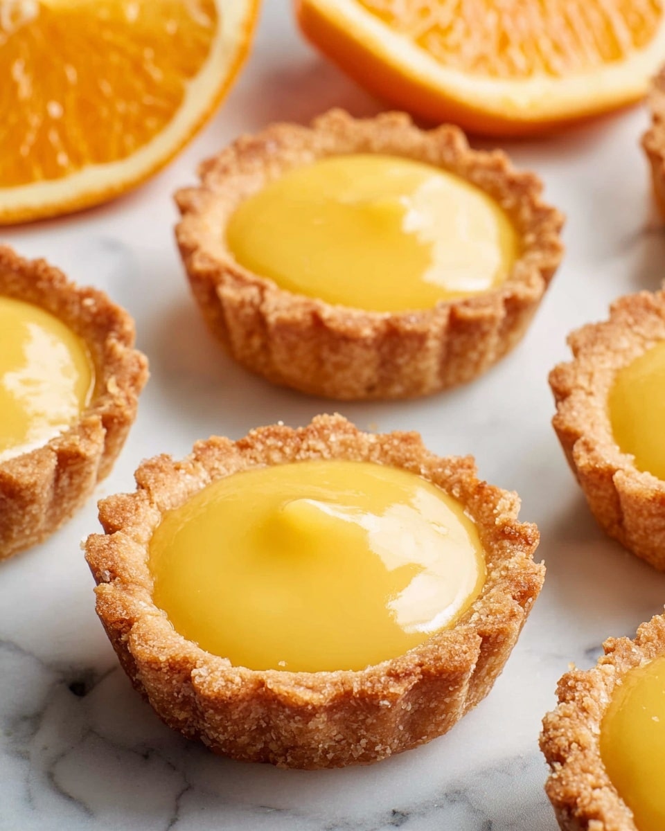 The image shows multiple small round tarts arranged on a white marbled surface. Each tart has two layers: a golden-brown, crumbly crust that forms a shallow cup with scalloped edges, and a smooth, glossy yellow filling that looks creamy and thick, filling the crust to the top without spilling over. In the background, there is a partially visible orange slice adding a fresh contrast to the warm tones of the tarts. The lighting highlights the glossy texture of the filling and the crumbly texture of the crust, making the tarts appear fresh and inviting. photo taken with an iphone --ar 4:5 --v 7
