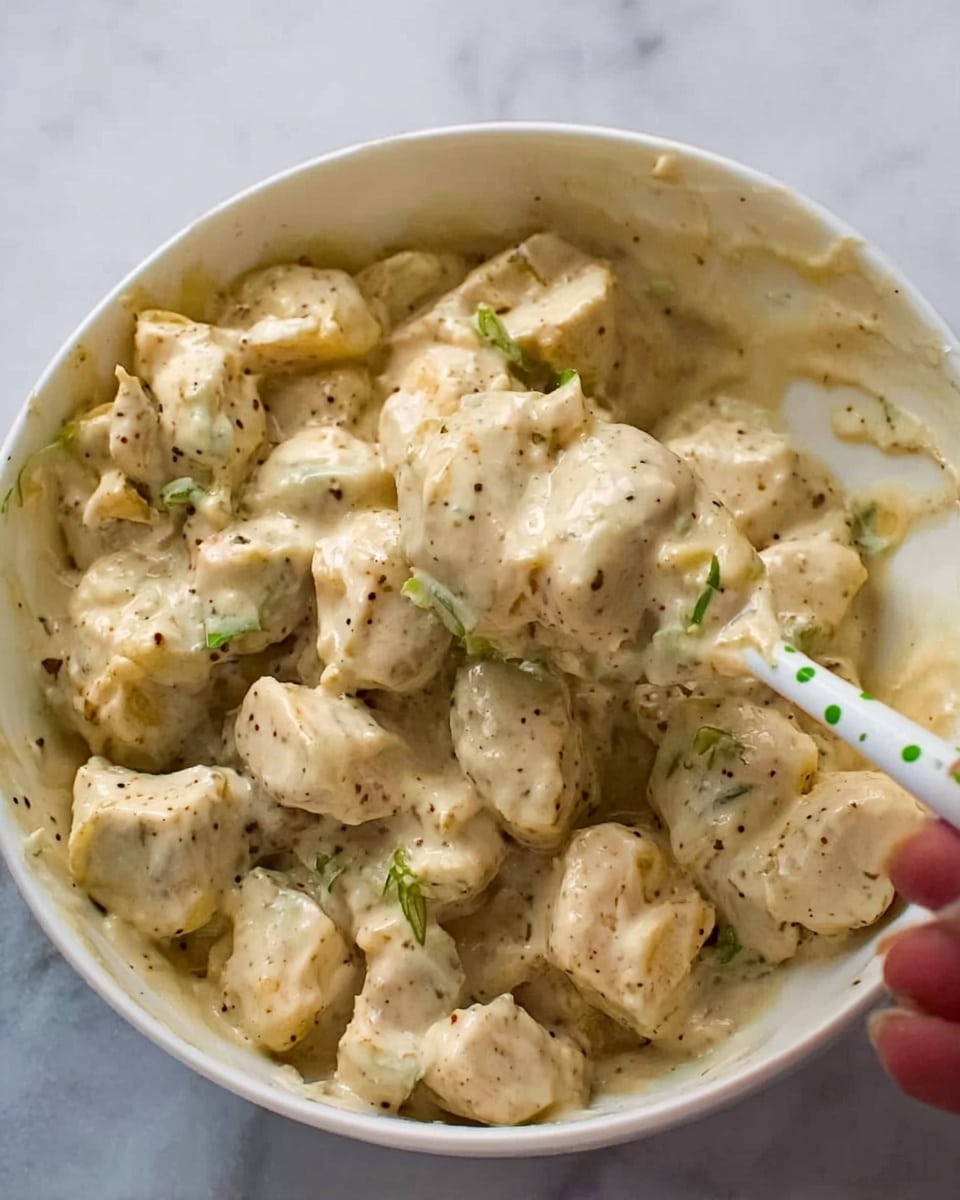 The image shows a white bowl filled with a creamy mixture of bite-sized pieces of light beige food, coated in a thick, pale sauce with visible small green and black seasoning flecks spread evenly. A woman's hand holding a white spoon with green dots is partially visible on the top right side, scooping the mixture. The bowl sits on a white marbled surface, adding a clean and bright background to the dish. The texture appears smooth and creamy with chunks clearly visible throughout, giving a rich but soft look. Photo taken with an iphone --ar 4:5 --v 7