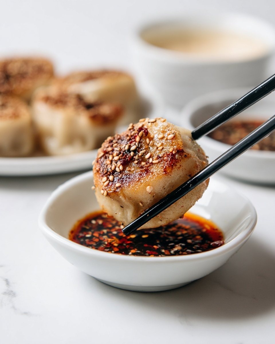 A small round dumpling with a golden-brown crispy outside covered in sesame seeds is held by black chopsticks. The dumpling has a textured top with some darker brown spots and is being dipped into a shallow white bowl filled with dark red soy sauce mixed with sesame seeds and red chili flakes. In the blurred background, there is a white plate with more dumplings and a white bowl with a creamy light-colored sauce, all placed on a white marbled surface. photo taken with an iphone --ar 4:5 --v 7