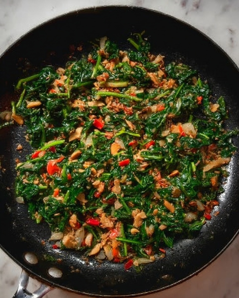 The image shows a black frying pan filled with a colorful mix of cooked ingredients. The dish has roughly three layers: the bottom layer is a dark skillet surface, the middle layer consists of green leafy spinach that looks soft and slightly wilted, scattered evenly across the pan; the top layer includes small pieces of light tan nuts or seeds, bright red bits that seem like chopped vegetables or peppers, and some light brown chunks mixed throughout, adding texture and color contrast. The whole dish looks lightly cooked and mixed well. The background is a white marbled surface. photo taken with an iphone --ar 4:5 --v 7