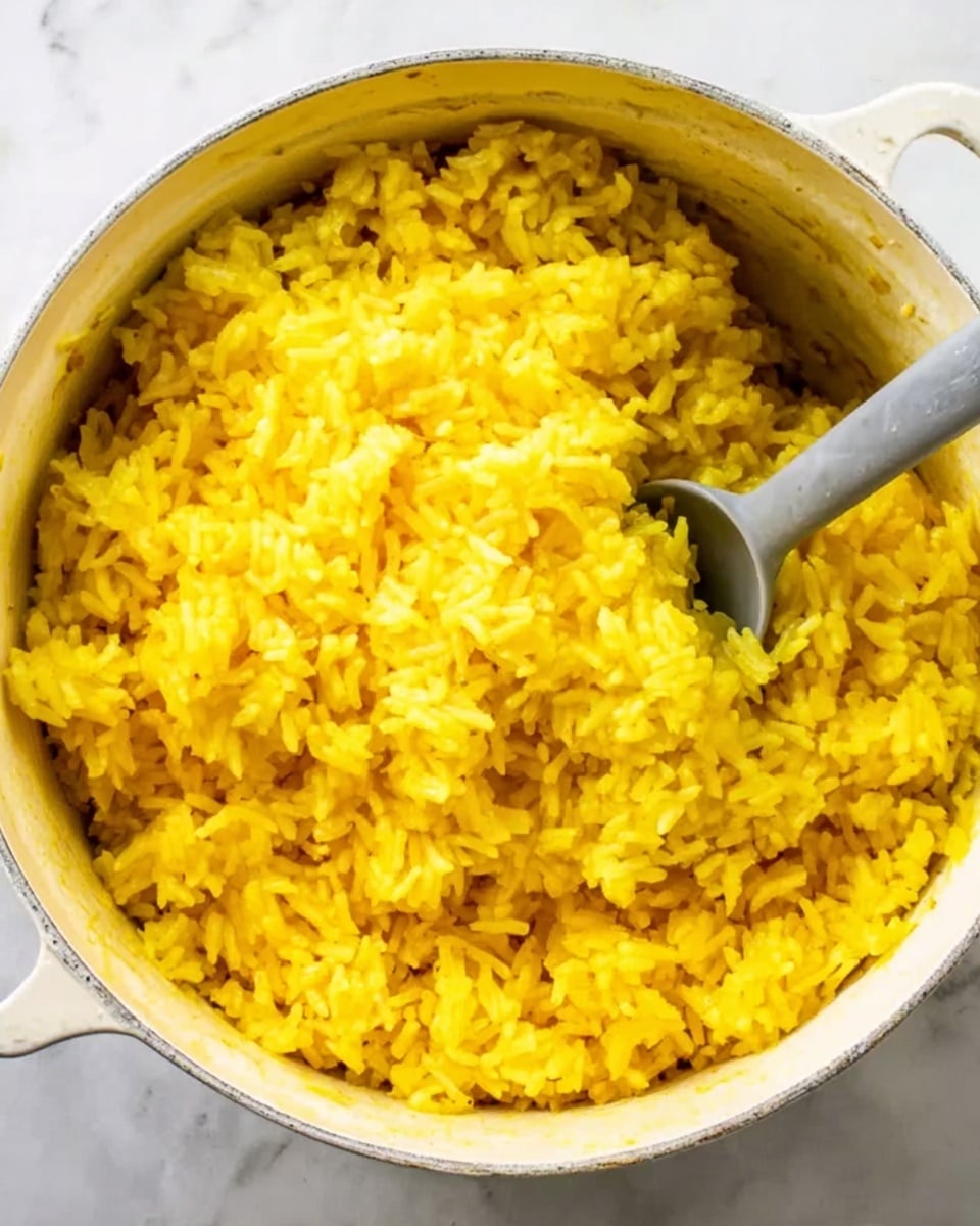 The image shows a close-up of a white pot filled with yellow rice that looks soft and fluffy. The rice grains are well-cooked and mixed evenly, with a shiny texture that suggests it is moist. Inside the pot, there is a gray spoon partially submerged in the rice, ready to serve. The background is a white marbled surface. The overall look is warm and inviting. photo taken with an iphone --ar 4:5 --v 7