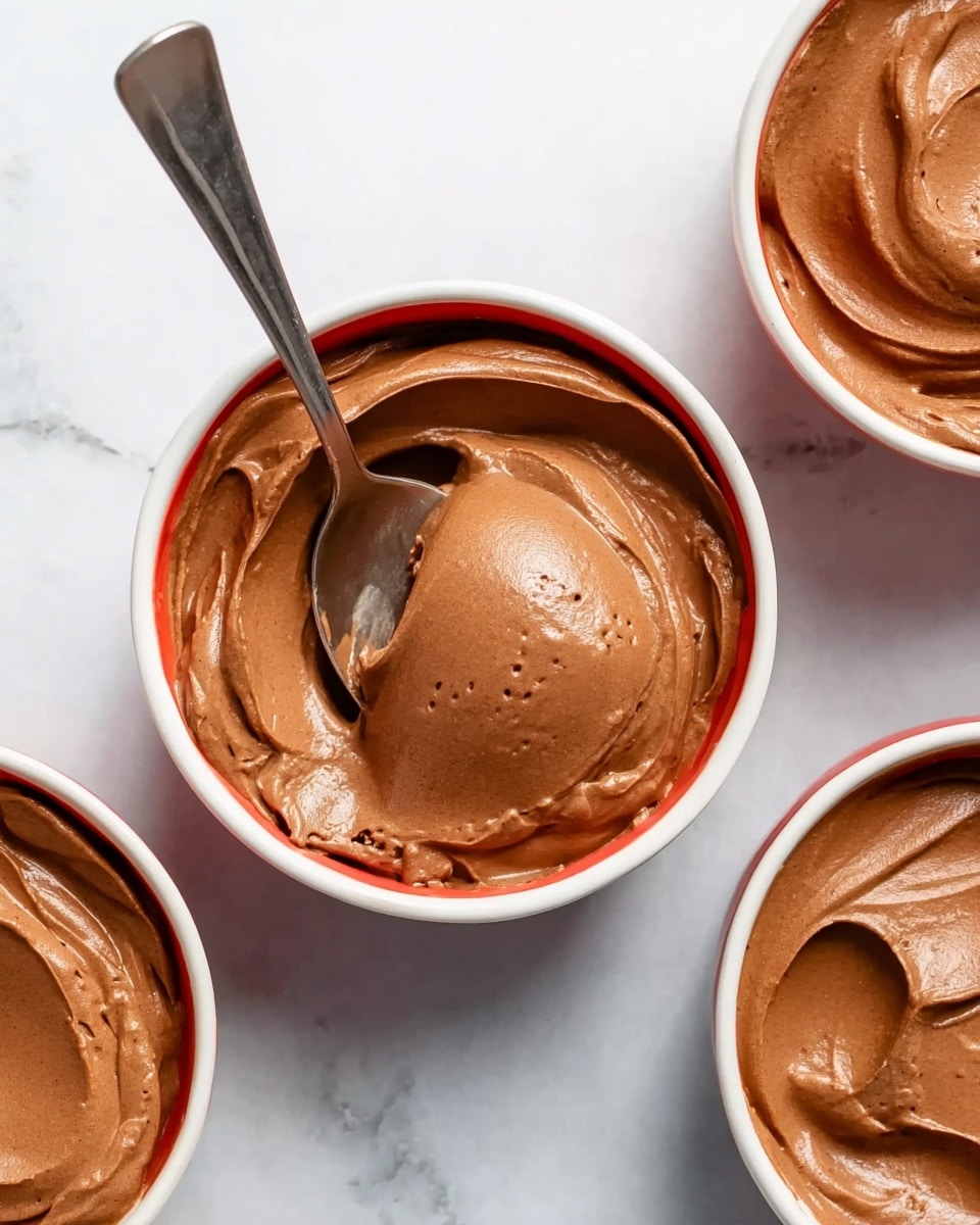 The image shows three white bowls filled with smooth, creamy chocolate mousse. The mousse in the center bowl has a large spoon scoop resting on the surface, revealing a light, airy texture with small air bubbles. The mousse's surface is shiny with soft waves and swirls. Each bowl sits on a white marbled surface with a silver spoon near each bowl, the spoon on the right half-covered with some mousse. The overall color is a rich brown of the mousse contrasting with the bright white bowls and background. photo taken with an iphone --ar 4:5 --v 7