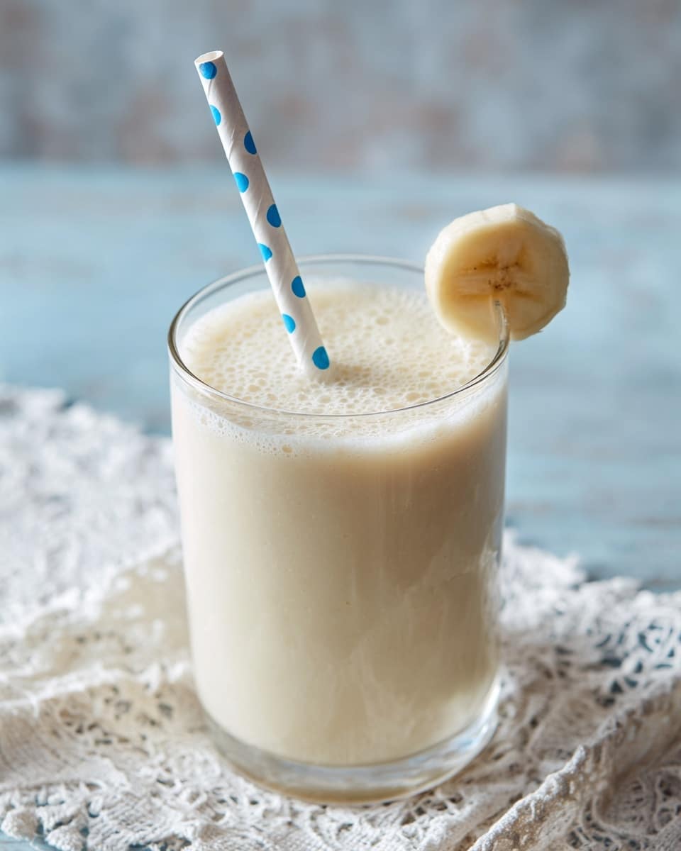 A creamy, light beige banana smoothie fills a clear glass, topped with tiny bubbles on the surface. A white paper straw with large blue dots stands upright inside the glass. A single round banana slice is placed on the rim of the glass. The glass is sitting on a delicate white lace cloth over a white marbled surface. The background is softly blurred with light blue and neutral tones. photo taken with an iphone --ar 4:5 --v 7
