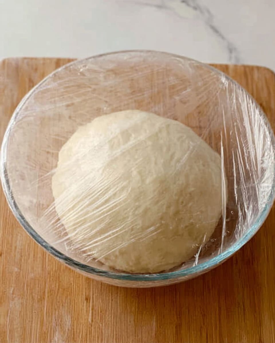 A clear glass bowl holds a smooth, round ball of pale dough with a soft texture, covered loosely by clear plastic wrap. The bowl sits on a light brown wooden surface, but the background is changed to a white marbled texture. The dough looks fresh and ready to be used, with a slight shine on its surface that suggests it is moist and well-kneaded. photo taken with an iphone --ar 4:5 --v 7