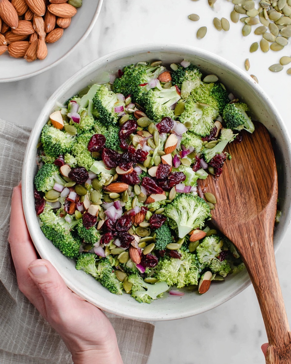 A white bowl filled with a colorful broccoli salad showing three main layers: chopped bright green broccoli pieces covering the bottom and sides, a mix of small diced red onion and dried red cranberries scattered on top, and whole almonds along with flat pale green pumpkin seeds spread over the salad. A wooden spoon is partly resting inside the bowl on the right side, and a woman's hand is holding the bowl at the bottom edge. The scene is set on a white marbled textured surface with a white plate containing almonds and pumpkin seeds visible in the top left corner of the image photo taken with an iphone --ar 4:5 --v 7