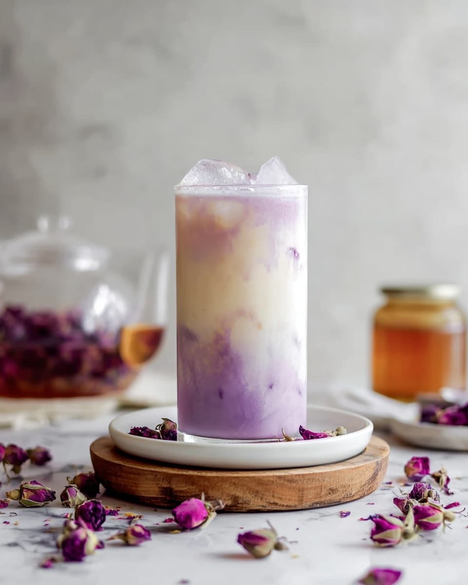 A tall clear glass filled with a three-layered drink stands on a white round plate, which is placed on a small wooden round board, all set on a white marbled surface. The drink’s bottom layer is a soft purple with a smooth texture, fading into a middle cream-white layer that blends gently upwards. The top layer is creamy white with purple streaks flowing down into the middle, topped with several ice cubes that give a slightly frosty look. Around the plate and surface, there are scattered dry purple rose petals and dried rosebuds, adding a delicate touch. In the background, a glass teapot with more purple petals and a small amber jar appear softly blurred. The photo taken with an iphone --ar 4:5 --v 7