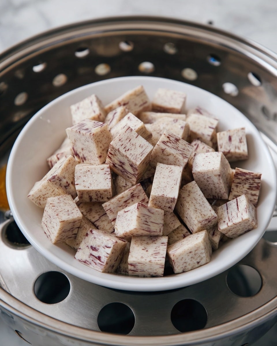 A white bowl filled with two layers of cubed taro root. Each cube is light beige with fine dark purple streaks running through it. The bowl is sitting inside a silver metal steamer pot with round holes visible on the bottom tray. The background is a white marbled surface. photo taken with an iphone --ar 4:5 --v 7