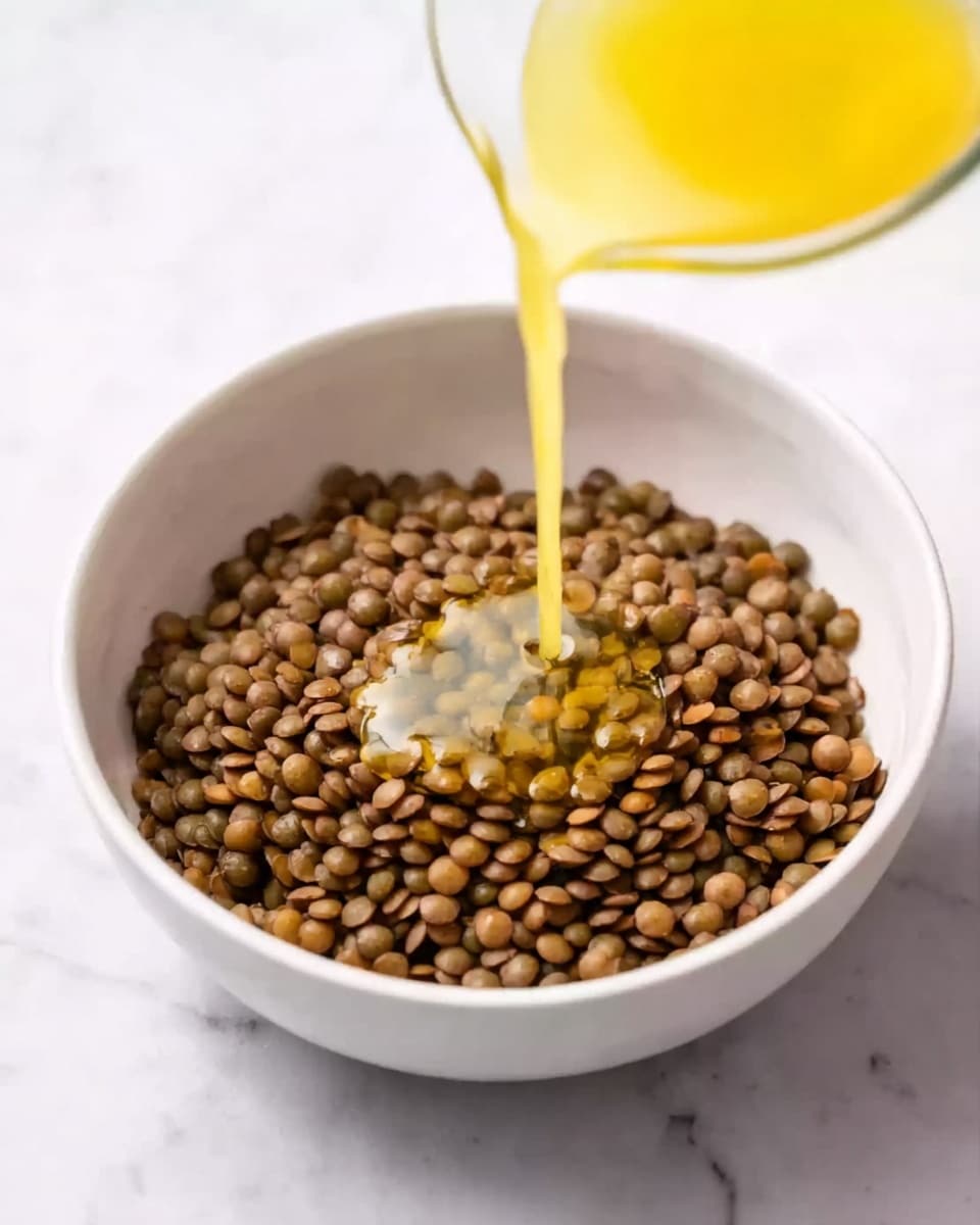 A white bowl filled with a single layer of small, round, brown lentils. A light yellow, smooth liquid is being poured into the center of the lentils from above, forming a small pool that slightly spreads on the lentils. The bowl is placed on a white marbled surface. Photo taken with an iphone --ar 4:5 --v 7