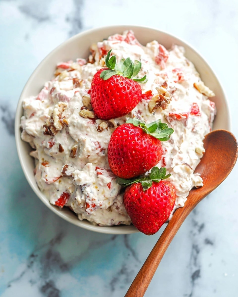 A white bowl filled with a creamy mixture that has small pieces of red strawberries and light brown nuts throughout. On top, three whole bright red strawberries with green leaves are placed in a row. A wooden spoon rests inside the bowl on one side, and the bowl sits on a white marbled surface. The creamy mixture looks thick and textured with visible chunks of fruits and nuts photo taken with an iphone --ar 4:5 --v 7