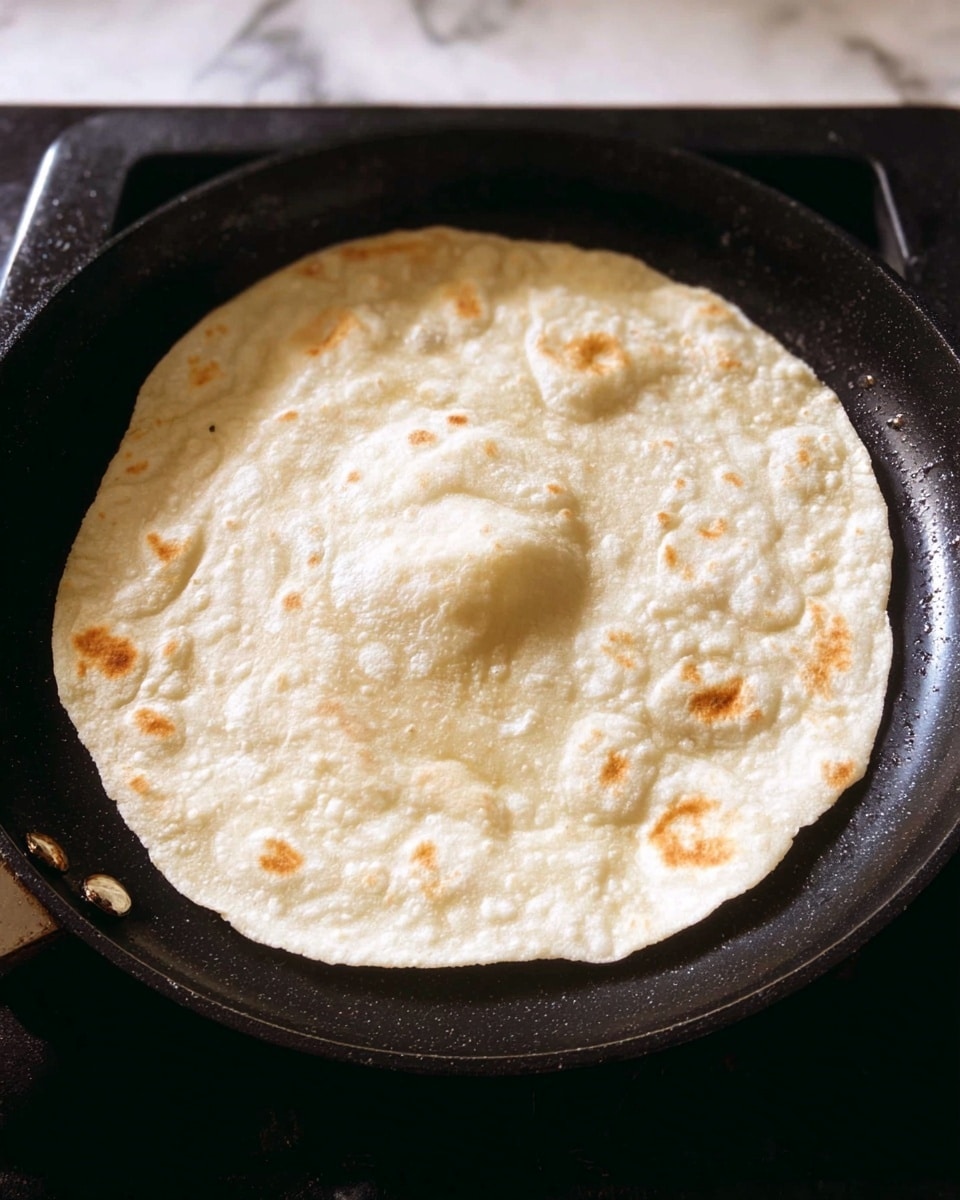 A close-up view of a single round tortilla cooking on a black pan over a stove, showing small bubbles on its surface with one larger puff in the center. The tortilla is light cream color with slight golden brown spots and a soft, slightly uneven texture. The pan contrasts with the white marbled background visible behind it. photo taken with an iphone --ar 4:5 --v 7