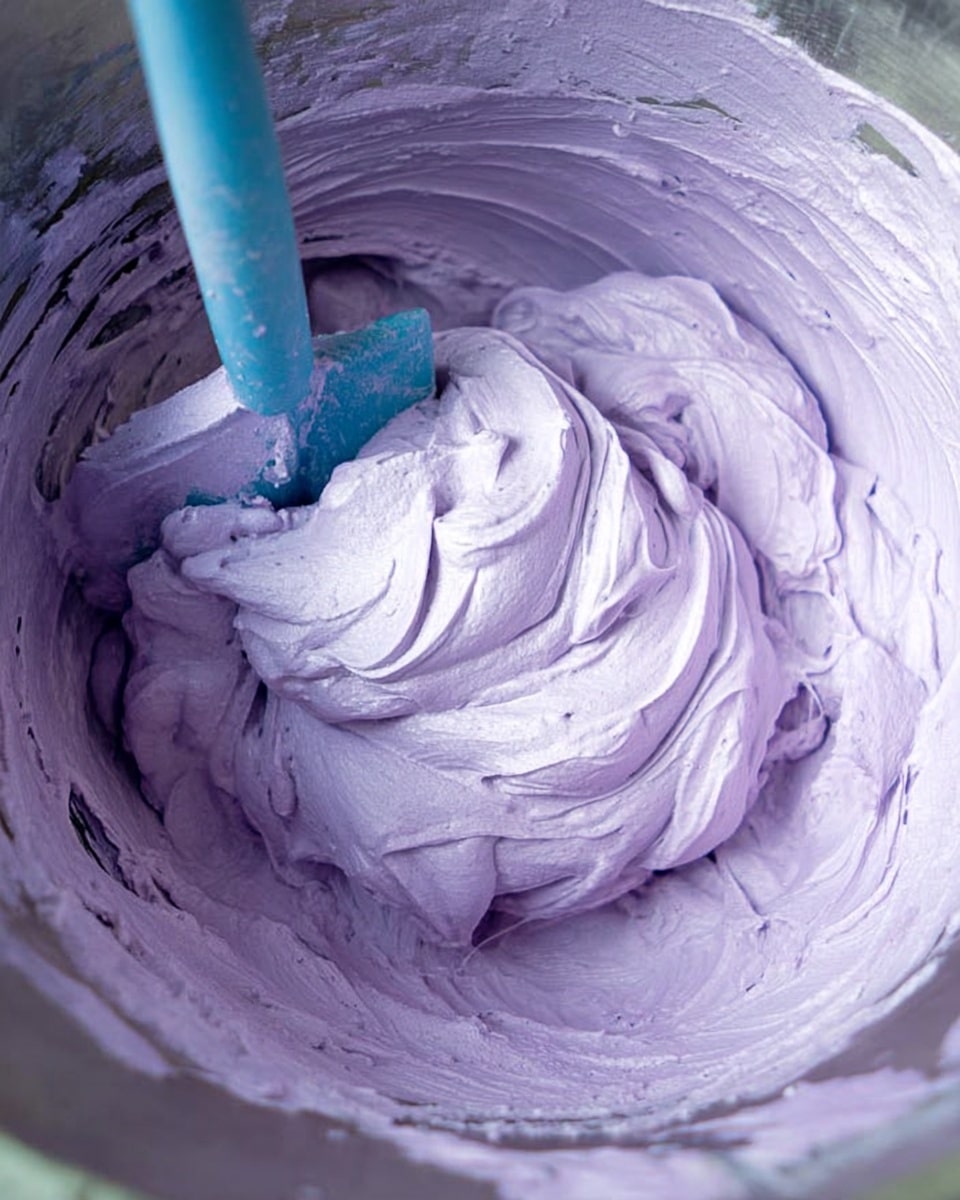 The image shows a close-up view of a light purple whipped mixture inside a metal bowl. The mixture is fluffy and smooth with soft peaks and swirls all around. A bright blue spatula is partly inserted into the mixture, creating gentle ridges as it moves through the creamy texture. The metal bowl has a shiny surface that reflects some of the purple color. The background is a white marbled texture. Photo taken with an iphone --ar 4:5 --v 7