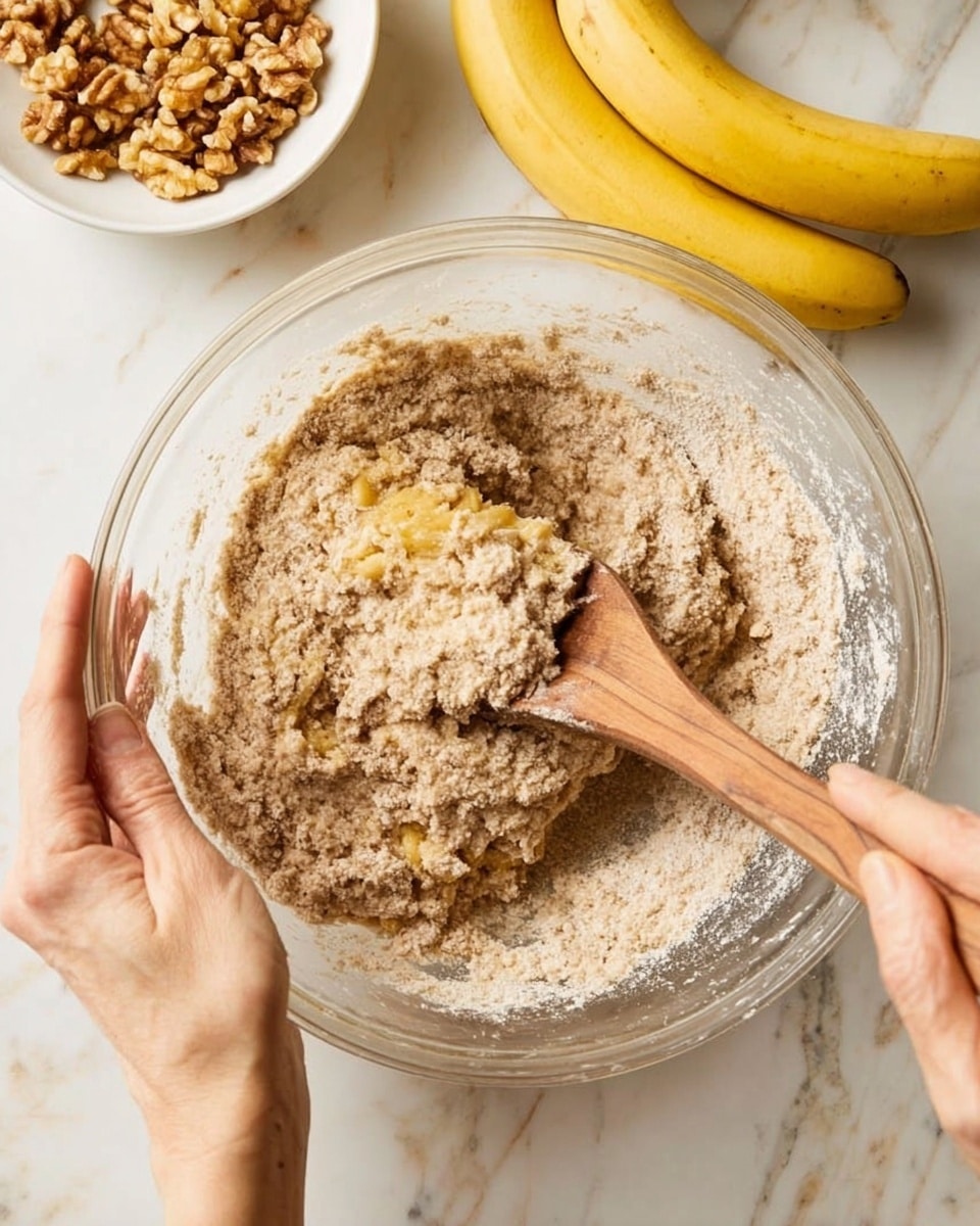 A clear glass bowl sits on a white marbled surface, filled with a mixture of light brown flour and mashed yellow bananas being stirred with a wooden spoon. A woman's hand holds the bowl steady on the left side, while another woman's hand uses the spoon to mix the ingredients, creating a textured, chunky batter. In the background, two ripe yellow bananas rest near the bowl, and a white bowl filled with chopped walnuts is placed slightly behind the mixing bowl. The overall scene shows a close-up, natural light view of the baking preparation. photo taken with an iphone --ar 4:5 --v 7