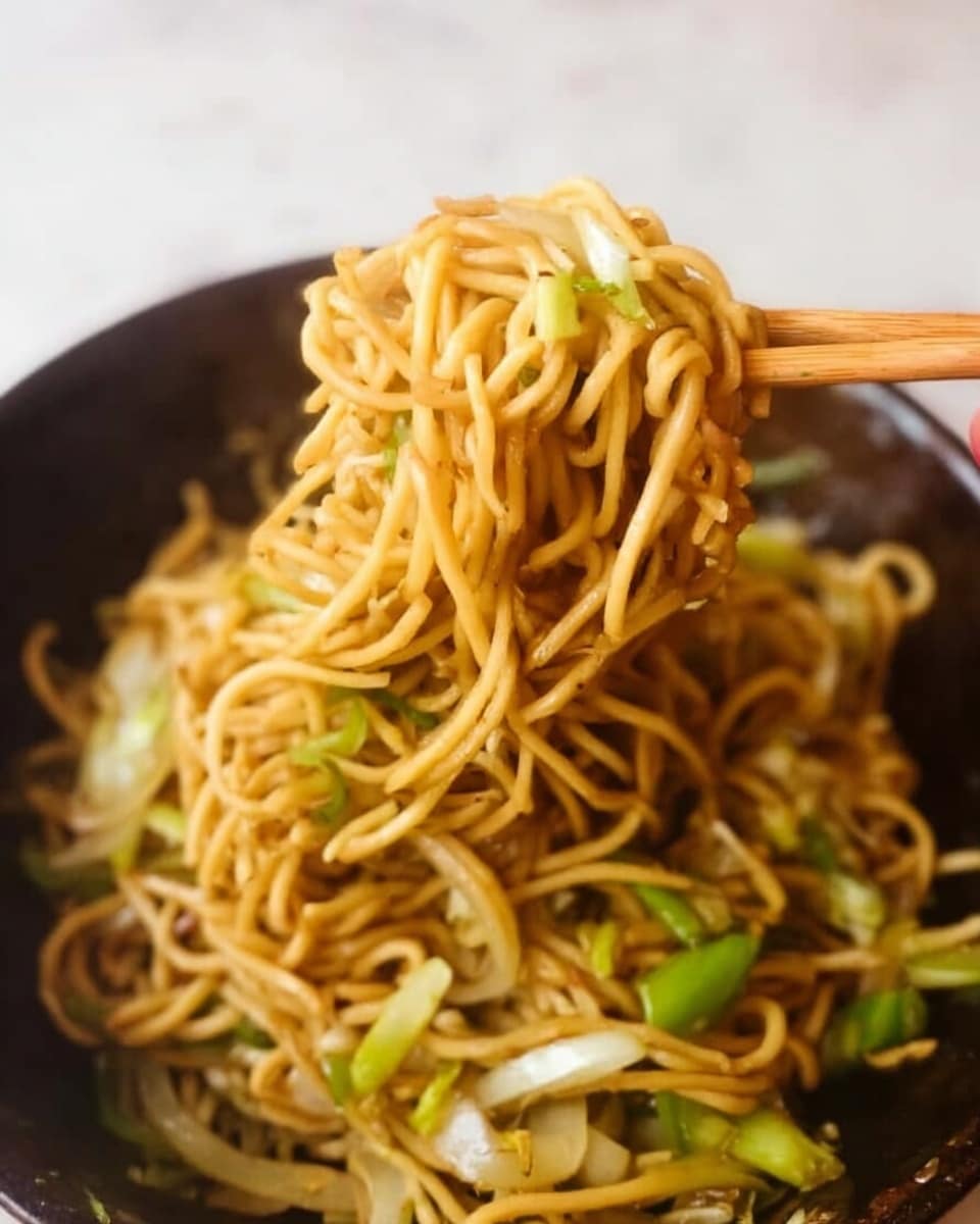 A close-up image of a serving of cooked yellow noodles mixed with light green sliced vegetables and small pieces of white onion. The noodles look soft and slightly shiny with a light brown sauce coating them. A pair of light wooden chopsticks, held by a woman's hand, lifts a portion of the noodles above a dark bowl, showing the noodles tangled and some vegetables sticking out. The background is a white marbled surface. photo taken with an iphone --ar 4:5 --v 7