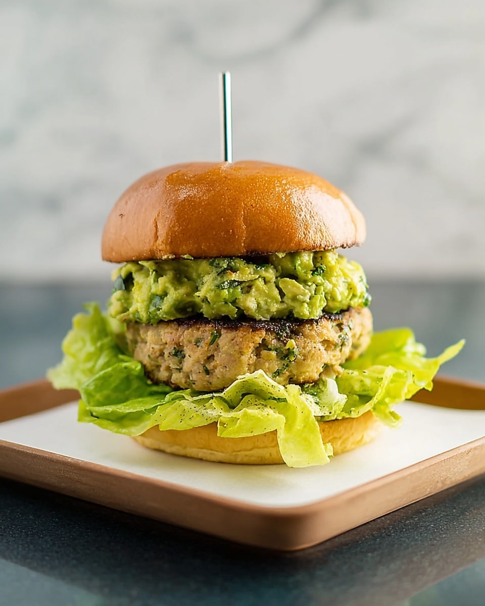 A burger with four main layers sits on a white square plate with rounded edges on a white marbled surface. The bottom layer is bright green, ruffled leafy lettuce. Above it is a thick, light brown grilled patty with slight char marks. On top of the patty is a chunky, bright green guacamole layer with visible bits of avocado and herbs. The burger is topped with a shiny, golden-brown soft bun. A small metal skewer is stuck in the middle from top to bottom. The background is plain and blurred to focus on the burger. Photo taken with an iphone --ar 4:5 --v 7