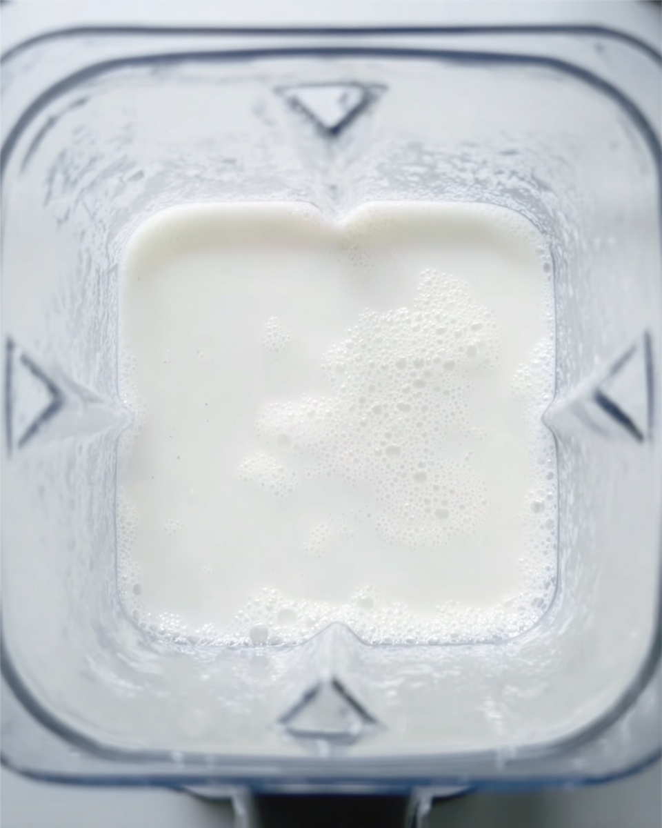 The image shows a close-up top view of a blender filled with a smooth, white liquid. The liquid layer fills most of the blender's clear container, with a slightly frothy texture around the edges. The blender container has a square shape with rounded corners, and the white marbled surface can be seen faintly around the base outside the container. The lighting is bright and natural, highlighting the clean and fresh appearance of the liquid. photo taken with an iphone --ar 4:5 --v 7