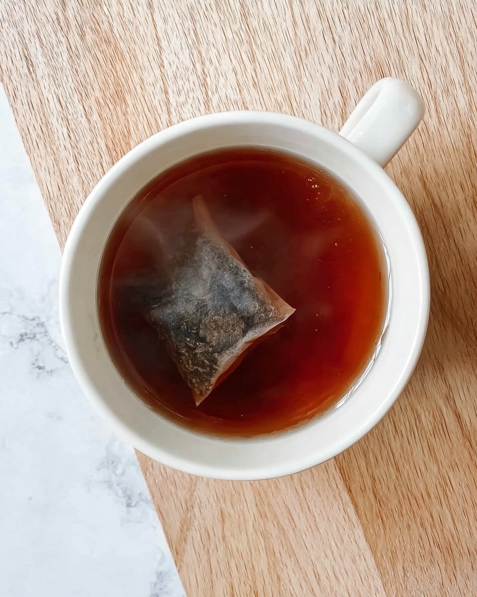 A white cup filled with dark brown tea, showing a steeped tea bag inside, the tea bag is partially transparent revealing tea leaves, the cup sits on a light wood textured surface which contrasts with the white marbled background, the tea looks warm with steam faintly visible above the cup photo taken with an iphone --ar 4:5 --v 7