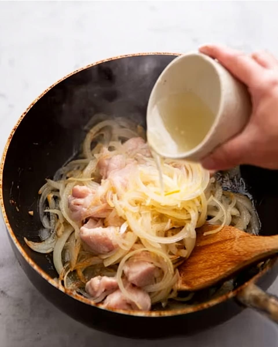 A close-up image shows a dark cooking pan filled with thinly sliced yellow onions that have a soft, translucent texture, arranged mainly around the edges, while raw pieces of light pink chicken sit in the center. A woman's hand is holding a small white bowl, pouring a light liquid into the pan, and there is a wooden spatula resting inside the pan, partially stirring the contents. The surface beneath the pan is white with a marbled texture. Steam rises gently from the pan, adding a warm and fresh feeling to the scene. Photo taken with an iphone --ar 4:5 --v 7