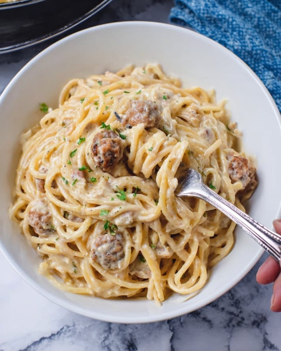 A white bowl filled with creamy spaghetti pasta, coated evenly in a light beige sauce with visible small chunks of sausage pieces throughout. The pasta strands are twisted in loose spirals, with a few sausage pieces sitting on top, showing a slightly browned texture. Tiny green herb bits are scattered lightly over the dish, adding a touch of color. A silver fork, held by a woman's hand, lifts a small portion of the spaghetti and sausage. The bowl rests on a white marbled surface with part of a black pan and a blue cloth visible in the background. photo taken with an iphone --ar 4:5 --v 7