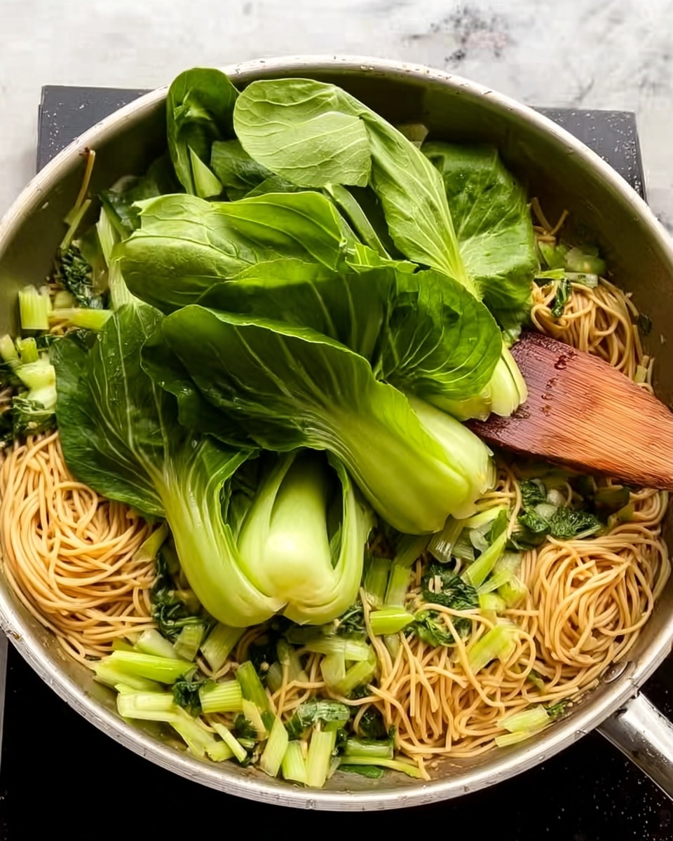 A metal pan filled with cooked yellow noodles at the bottom, mixed with light green chopped vegetables including stalks and leaves. On top, several whole bright green large leaves of bok choy are added, covering a large part of the noodles. A wooden spatula rests on the right side of the pan, partly touching the noodles and vegetables. The background is a white marbled texture. Photo taken with an iphone --ar 4:5 --v 7