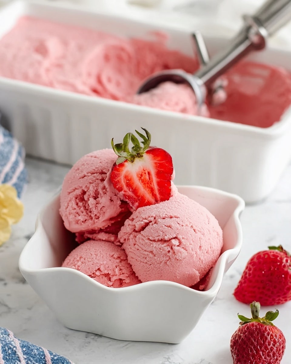 The image shows three scoops of pink strawberry ice cream with a smooth but slightly textured surface inside a white, wavy-edged square bowl. On top of the middle scoop, there is a half strawberry with visible seeds and a green leafy top, placed upright to show its red inside. Behind the bowl, there is a white rectangular dish filled with the same pink ice cream, with a metal scoop resting inside it and a whole strawberry placed near the scoop handle. The background has a white marbled texture with a small part of a blue and white striped cloth visible on the side. photo taken with an iphone --ar 4:5 --v 7