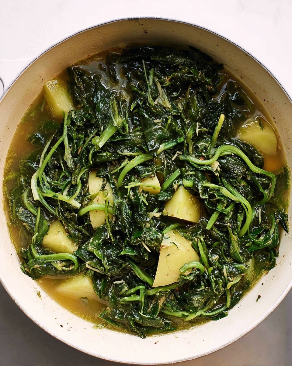This image shows a white pot filled with a cooked green leafy vegetable mixed with light yellow potato pieces. The greens are dark green with a wet, cooked texture, featuring visible stems and curling leaves. The potato pieces are scattered evenly throughout the greens and are soft with a smooth texture. A light brown broth covers the bottom part of the pot, partially soaking the vegetable and potatoes. The inside of the pot is white, and it sits on a white marbled surface. photo taken with an iphone --ar 4:5 --v 7