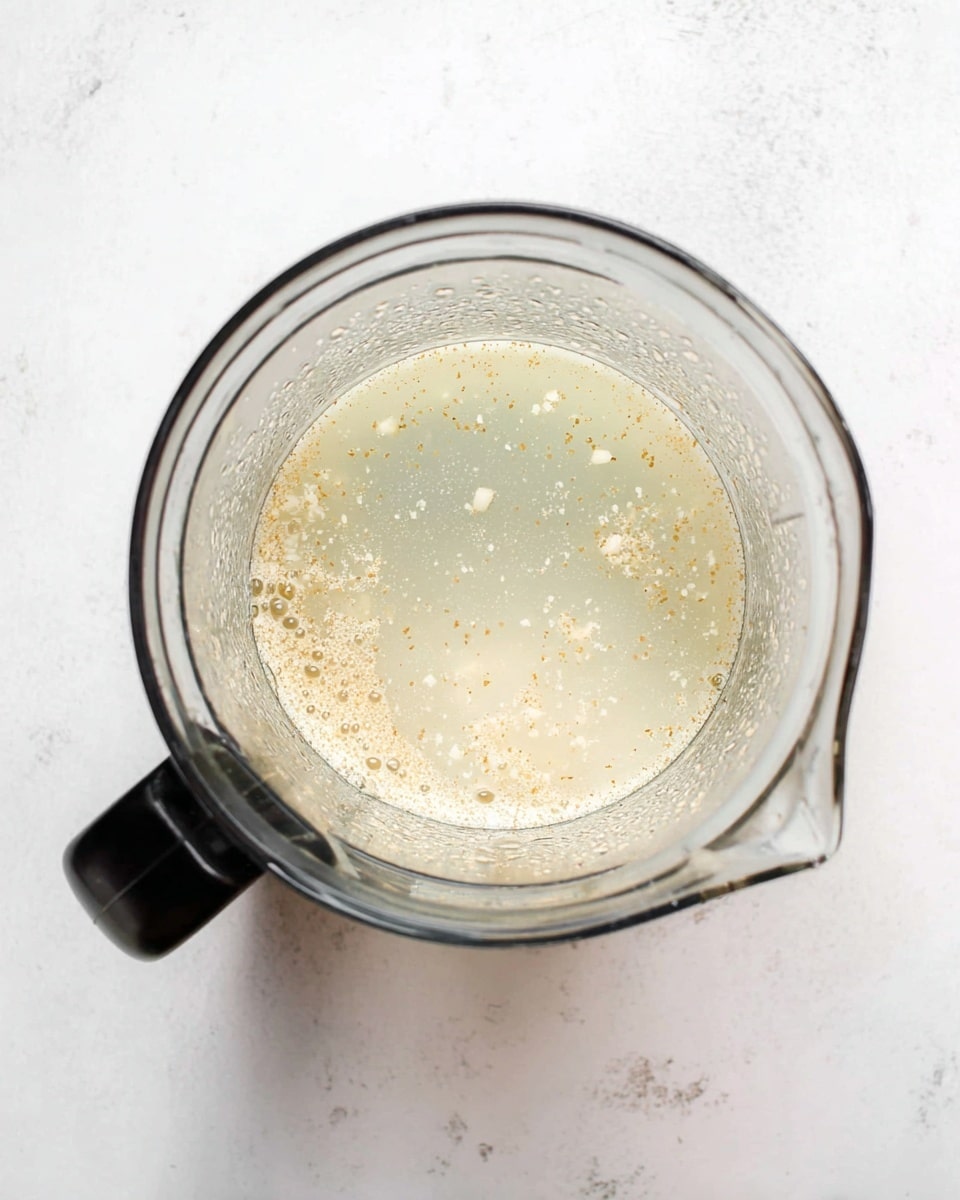 A top view of a clear blender container partially filled with a cloudy, pale yellow liquid with small floating bits, resting on a white marbled surface. The container has a black handle positioned to the bottom left. Inside, the liquid appears slightly textured with tiny specks spread evenly throughout. Photo taken with an iphone --ar 4:5 --v 7