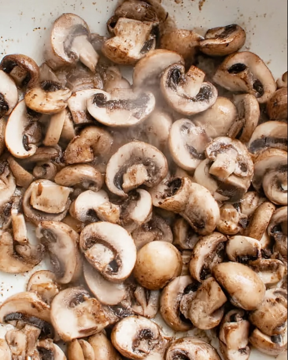 The image shows a close-up of many cooked mushrooms in a white shallow pan. The mushrooms are sliced into halves and quarters, with light brown caps and creamy white insides. They have a slightly shiny and moist look from being cooked, with some areas showing darker brown spots from browning. The mushrooms cover the entire white pan surface, giving a dense and textured appearance. The background is a white marbled surface. photo taken with an iphone --ar 4:5 --v 7