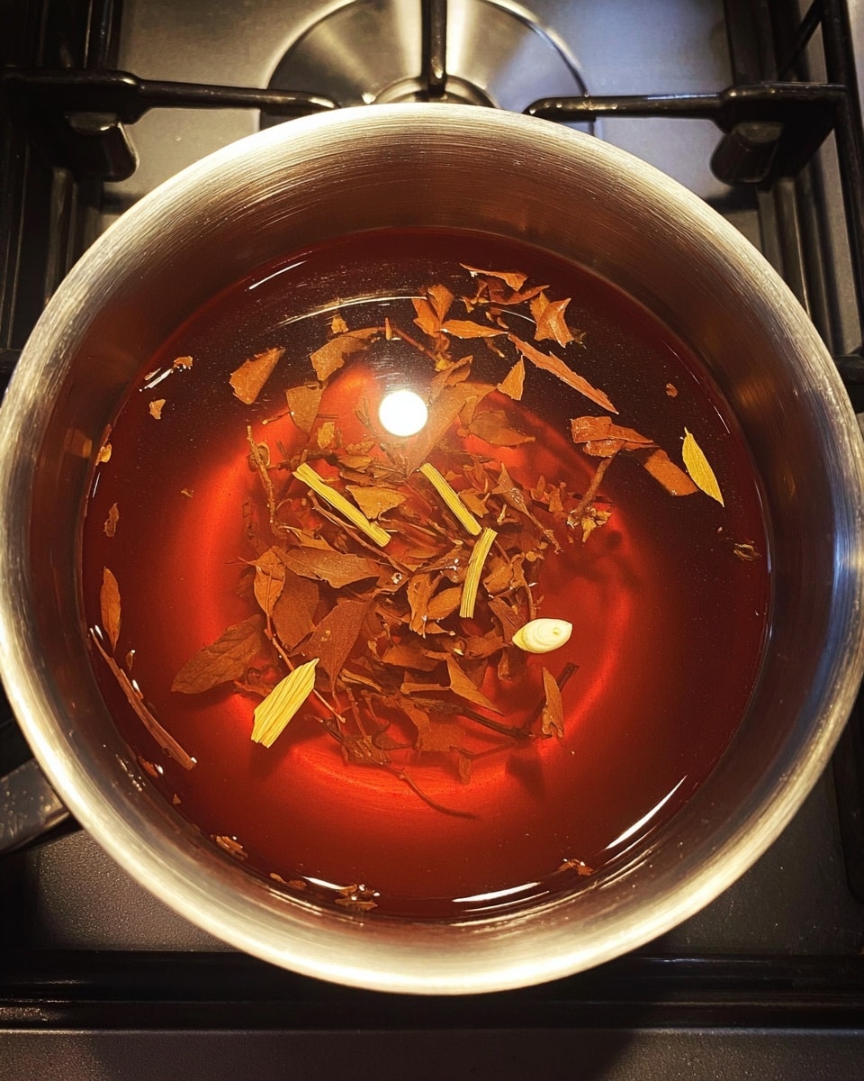 A shiny silver pot filled with clear reddish-brown liquid with small pieces of dried brown leaves and thin light yellow sticks floating on the top. A single small white clove is also visible near the edge inside the liquid. The pot is set on a stove with a black burner and silver control panel partially visible. photo taken with an iphone --ar 4:5 --v 7