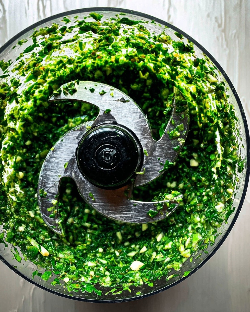 Top view of a food processor bowl filled with finely chopped green herbs mixed with small white bits, likely garlic or nuts. The sharp silver blades sit in the center, surrounded by the bright green and white mixture that clings to the sides of the clear bowl. The background is a white marbled texture surface. photo taken with an iphone --ar 4:5 --v 7